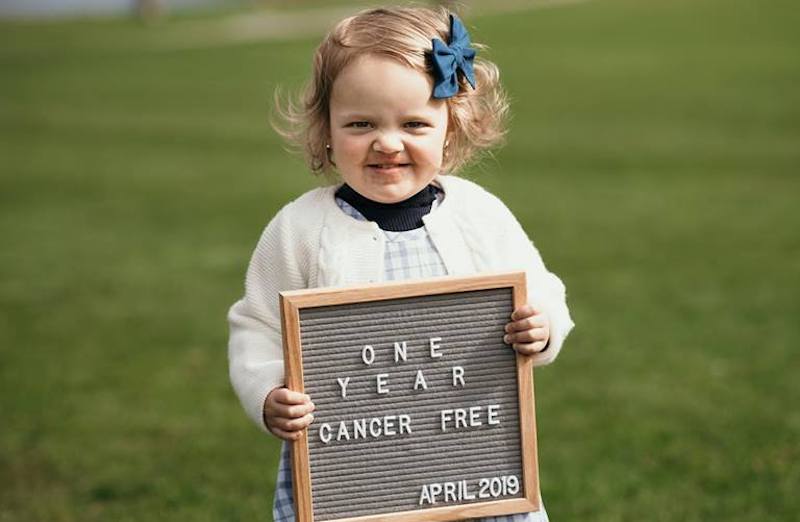 Stella, who was treated for neuroblastoma, holds a sign saying "One year cancer free."