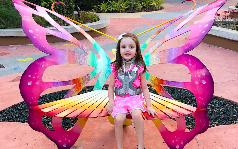 Gracie, wearing a brace after spinal fusion surgery, sits on a bright, pink butterfly bench. 