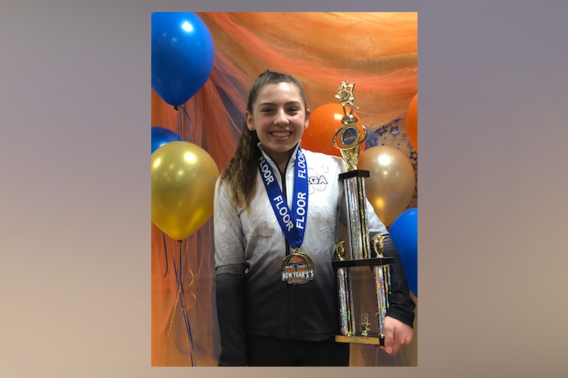 Mikayla, who was treated for hip avulsion fracture, smiles with two medals around her neck and a large trophy in her arms.