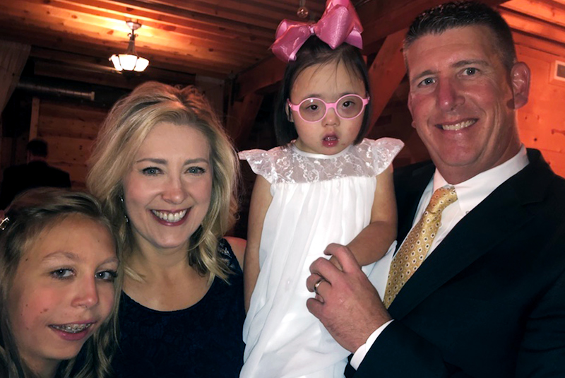 The Gottschalks pose for a photo at a wedding. Everli wears a white, flower girl dress. 