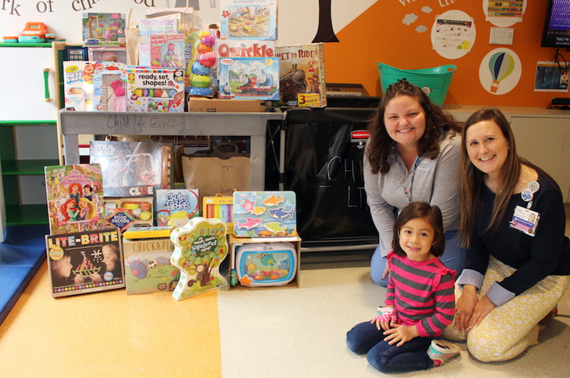 Kalea, who had moyamoya surgery, poses with her clinicians in front of  toys she raised for a playroom at the hospital