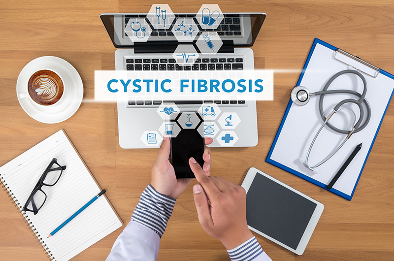 Cystic fibrosis banner is centered in front of a laptop and surrounded by tiny icons representing different health aspects of the condition, also with the hands of a person typing on mobile phone, and an iPad nearby.