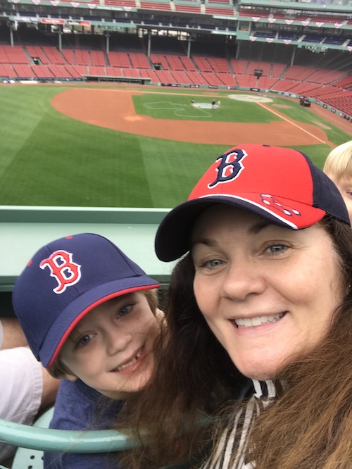 Mom and her young son wearing baseball hats sitting in the stands at Fenway Park in front of the baseball field for a game.
