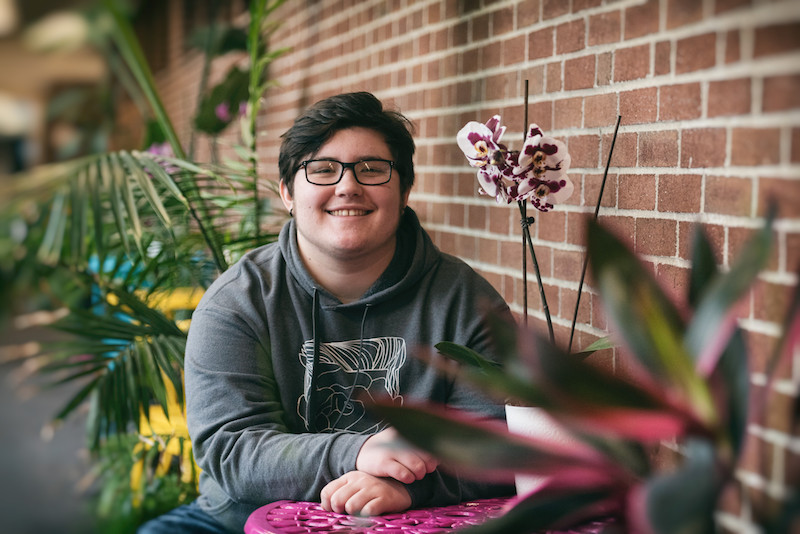 Oliver, a trans teen, sits amid tropical flowers and smiles at the camera.