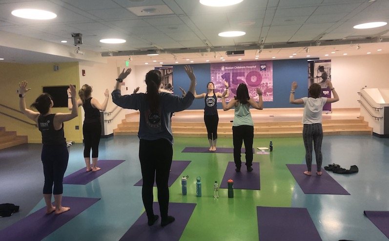 A female yoga instructor stands with her feet together and demonstrates holding her hands up in a pose in the front of a big room and a group of young women with IBD copy her movements.