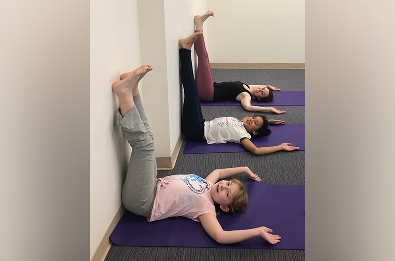 Two young girls with IBD lie on yoga mats on the floor next to an instructor as they all stretch their legs up along the wall.