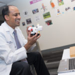 Doctor holds up toy cow as he has a telemedicine visit on his computer