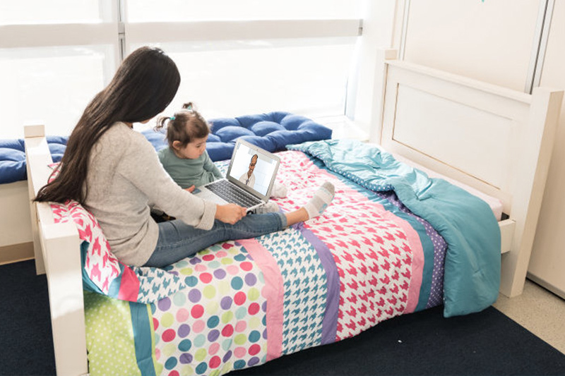 A woman and toddler sit on a bed with computer demonstrating a virtual visit