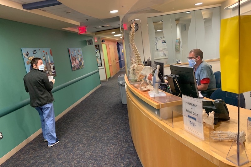 A clinician in the orthopedic urgent care unit sits at a desk behind plexiglass as another walks past. Both are also wearing masks to prevent the spread of COVID-19. 