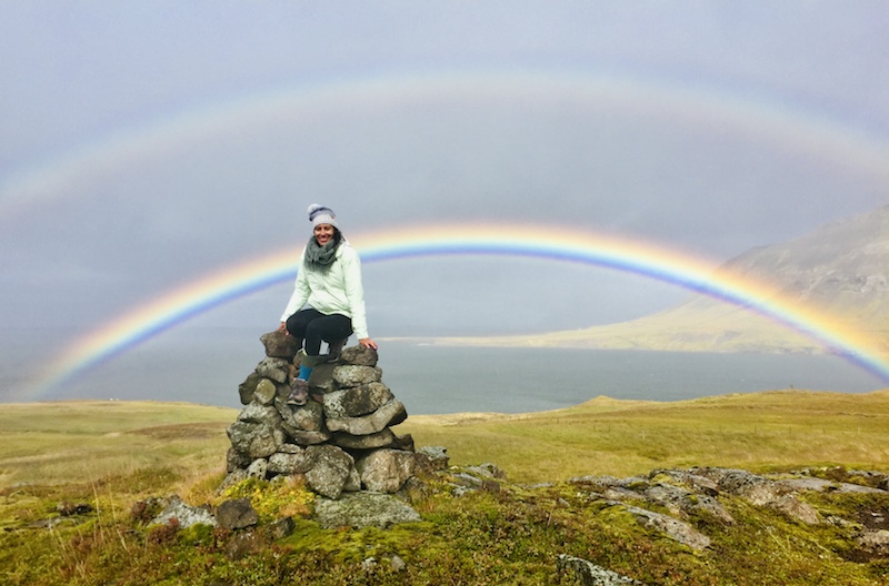 Carla Coutinho  in a hat and scarf, poses on a rock structure with a double rainbow in the sky behind her. 