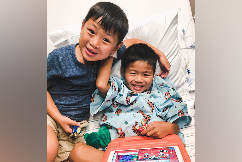 Steven in a hospital gown after surgery for anorectal malformation. His brother Bo has his arm around him and both are smiling.