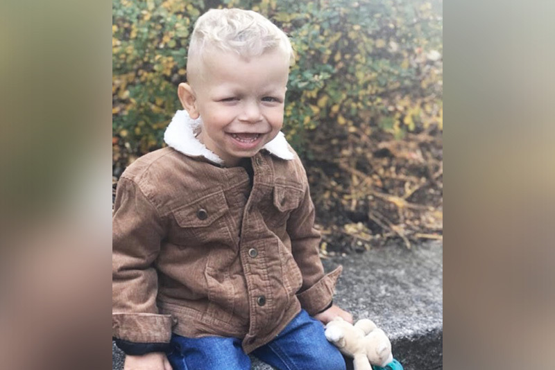 rylan, who has a laryngeal cleft, sits outside. he is wearing a brown jacket and smiling