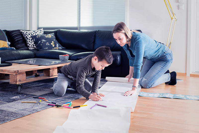 Boy and his mother drawing on a large sheet of paper in their living room.