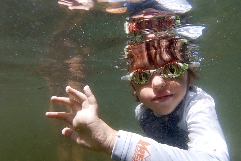 Colin, who was born with infantile scoliosis, looks at the camera underwater wearing lizard goggles.