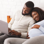 Pregnant woman and spouse lying on bed looking for pediatrician on computer