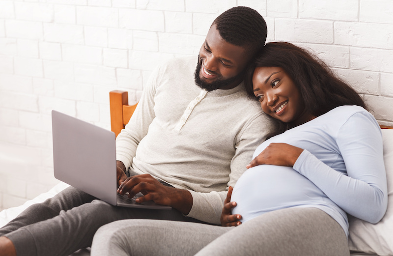 Pregnant woman and spouse lying on bed looking for pediatrician on computer