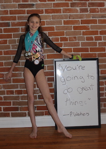 Ashley, who was born with symbrachydachtyly, in her gymnastic uniform, poses next to a whiteboard that says, "You're going to do great things." 