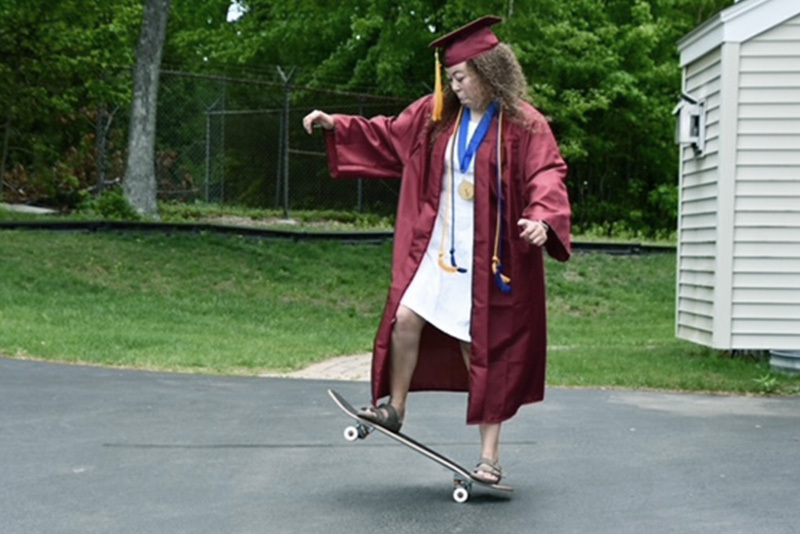 emily, who has graves disease, stands on her skateboard. she is wearing a graduation cap and gown