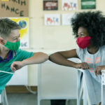 Two kids in a classroom wearing masks bump elbows across the aisle