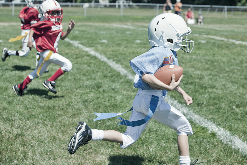 A young football player runs the ball down the field in a game of flag football. Sports played outdoors have a lower risk of COVID-19 than those played indoors.