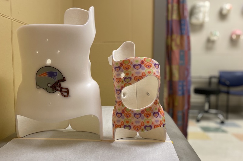 An adolescent scoliosis brace next to an infant scoliosis brace. Families can choose a color or pattern for the brace, which is tailor made for their child. In this case, the adolescent brace has a Patriots logo and the infantile brace is covered in pink hearts. 