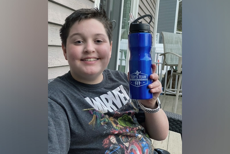 austin, who had two strokes, holds a water bottle. he is smiling and wearing a Marvel comics t-shirt