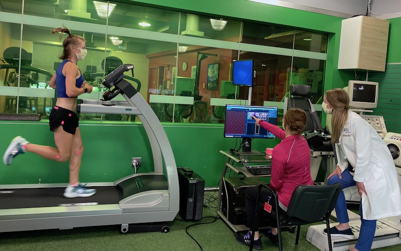 An athlete runs on a force-plate treadmill while sports medicine specialists review gait analysis data on a computer.