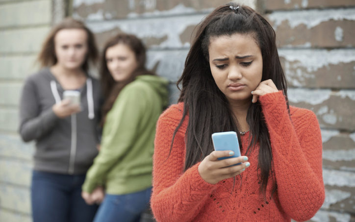 a teen girl looks sadly at her phone while two other girls laugh at her in a demonstration of bullying