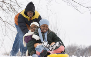 Two children sit in a snow tube as another child pushes them from behind.