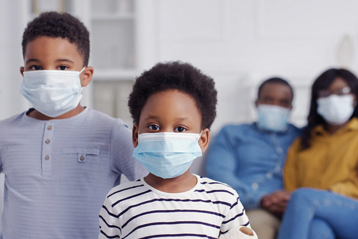 Two children wearing surgical masks with two adults in the background also wearing masks