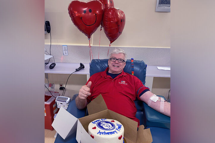 Frank donating blood with a cake on his lap. The cake is decorated with the Boston Children's logo and says "Happy Anniversary, Frank!" 
