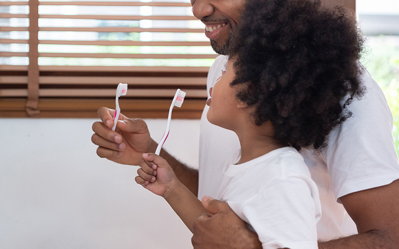 A parent and child, standing next to one another, both hold aloft toothbrushes.