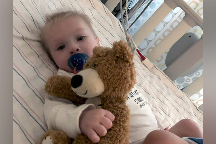 Infant Liam Allis clutches a brown stuffed bear while resting in a bed at Boston Children's Hospital.
