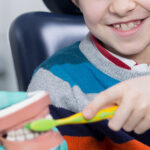 A child in a dentist chair, holding a toothbrush in their left hand, reaches over to a model of a mouth and brushes the teeth.