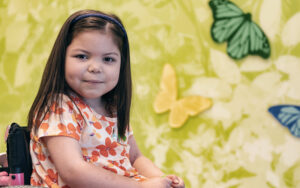 Kira, who had a kidney transplant, smiles next to a wall decorated with butterflies