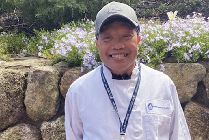 Willie Hernandez in a white chef's jacket, smiling outside with flowers in the background. 