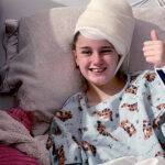 Young girl with bandaged head smiling and giving thumbs up from a hospital bed