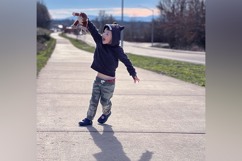 Levi Blair Winkler holds his stuffed monkey in the air as he playfully walks on a sidewalk.