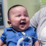 A smiling baby boy who who has a tracheostomy tube, sitting on a woman's lap.