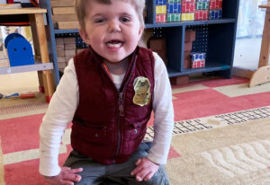 A young, toddler boy smiling on a playroom rug with his hands on his lap.