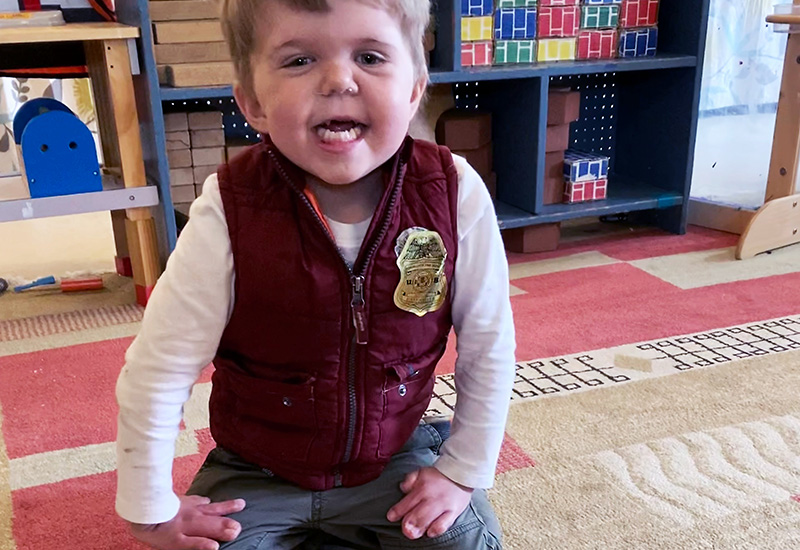 A young, toddler boy smiling on a playroom rug with his hands on his lap.