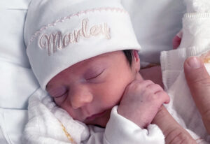 A sleeping newborn in a white cap grasping her father's finger.