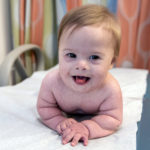 Baby Myles smiles on an observation table in the hospital, resting on his arms and smiling towards the camera.