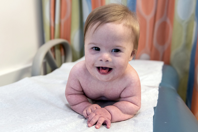 Baby Myles smiles on an observation table in the hospital, resting on his arms and smiling towards the camera.
