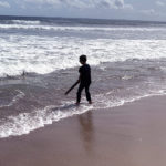The silhouettes of a little boy is standing on the shoreline of a beach holding a stick pointing down towards the water.