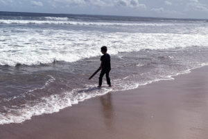 The silhouettes of a little boy is standing on the shoreline of a beach holding a stick pointing down towards the water.