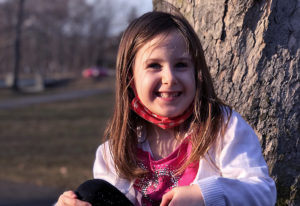 Macy smiles under a tree with her pink mask pulled down her chin.