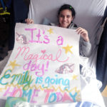 A young girl in a hospital bed holding a large paper sign that reads 
