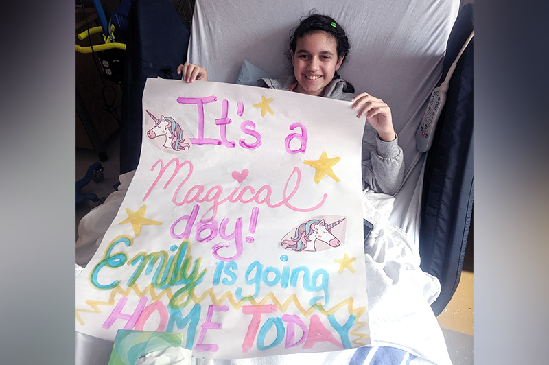 A young girl in a hospital bed holding a large paper sign that reads "Emily is going home today."