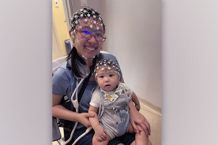 An infant in their smiling mother’s lap. Both are wearing EEG nets on their heads.